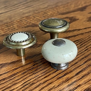 May include: Three decorative cabinet knobs with brass and ceramic details. One knob features a speckled, off-white ceramic top with a silver base. Another has a brass top and a beaded edge. The third knob has a brass top.