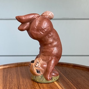 May include: A brown ceramic bunny figurine in a handstand pose. The bunny has a small, round tail and is standing on a patch of green grass. The figurine is on a wooden tray.