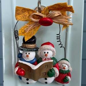 May include: A hanging ornament featuring three snowmen singing from a gold book with musical notes. The snowmen wear hats, scarves, and mittens in red, blue, green, and black. A yellow fabric bow and a red button are attached to the top wire hanger.