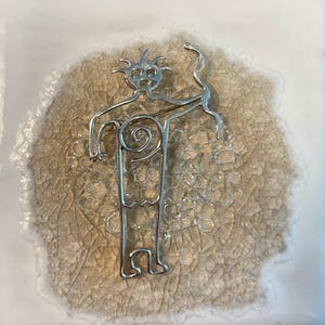 May include: A silver-colored metal figure with a stylized human form, featuring a head with radiating lines, a spiral torso, and outstretched arms. The figure is set against a textured, light brown background with a white border.