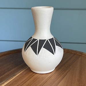 May include: A small, white ceramic vase with a geometric black design around the neck. The vase has a rounded base and a slender neck, standing on a wooden surface. The background is a light blue wall.
