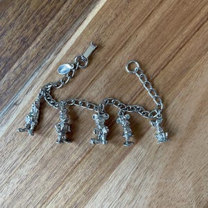 Op de afbeelding: Zilverkleurige bedelarmband met vijf karakterbedels. De armband heeft een schakelkettingontwerp en een sluiting. De bedels zijn gedetailleerde beeldjes van stripfiguren.