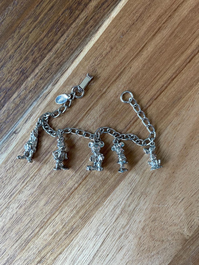 Op de afbeelding: Zilverkleurige bedelarmband met vijf karakterbedels. De armband heeft een schakelkettingontwerp en een sluiting. De bedels zijn gedetailleerde beeldjes van stripfiguren.