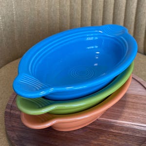 May include: A stack of three oval-shaped ceramic serving dishes in bright colors: blue, green, and orange. Each dish has a handle on either side and a concentric circle design in the center. The dishes are stacked on a wooden serving tray.