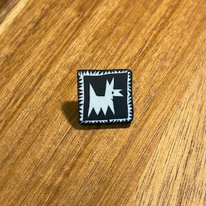 May include: A square pin featuring a black background with a white stylized dog illustration in the center. The dog is depicted with sharp, angular lines, and the entire square is framed by a white border with a jagged, sawtooth-like pattern.
