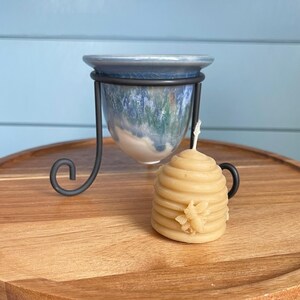May include: A ceramic bowl with a blue, green, and white mottled glaze sits in a black metal stand. Beside it is a light yellow, beehive-shaped beeswax candle featuring a small, molded bee detail on the front and a single white cotton wick.