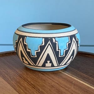 May include: A small, round ceramic bowl featuring a matte finish with a geometric pattern in black, cream, and bright turquoise. The design consists of alternating triangular and stepped shapes encircling the exterior, with a solid black rim.