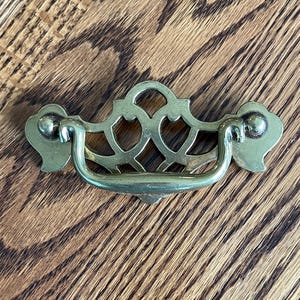 May include: A brass drawer pull with an ornate, openwork design. The pull has a curved handle and decorative end pieces. The hardware is a warm gold color, and is set against a wood grain background.