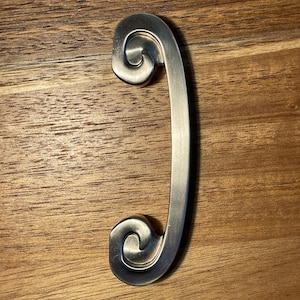 May include: A brushed silver metal cabinet pull featuring a curved, elongated handle with decorative scroll-shaped ends. The hardware has a smooth, matte finish and a symmetrical design, providing a functional grip for drawers or cabinet doors.