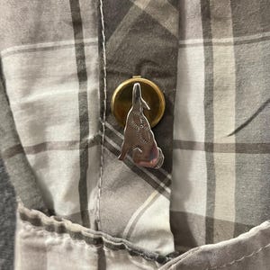 May include: A silver-colored pin in the shape of a howling wolf is attached to a button on a gray and white plaid shirt. The pin is mounted on a gold-colored circular base. The shirt has a pocket on the left side.
