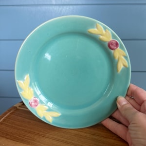 May include: A round ceramic plate with a smooth turquoise glaze. The rim features two raised, molded floral motifs, each consisting of a small pink circular center surrounded by four pointed yellow leaves, positioned on opposite sides of the plate.