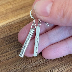 May include: A pair of silver dangle earrings. One earring features a series of etched triangles, while the other has a series of etched symbols. The earrings are suspended from a silver hook.