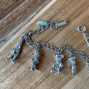 Op de afbeelding: Een zilverkleurige bedelarmband met Disney-personages. De armband bevat bedels van Mickey Mouse, Minnie Mouse, Donald Duck en andere figuren. De sluiting is gemarkeerd met het Disney-logo. De armband wordt gepresenteerd op een houten oppervlak.