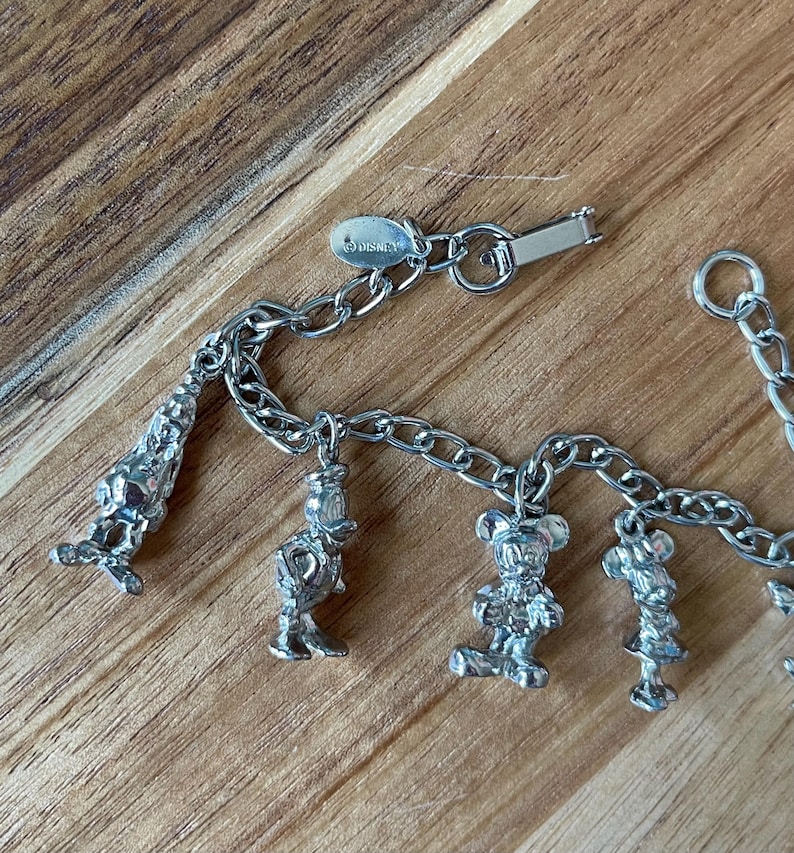 Op de afbeelding: Een zilverkleurige bedelarmband met Disney-personages. De armband bevat bedels van Mickey Mouse, Minnie Mouse, Donald Duck en andere figuren. De sluiting is gemarkeerd met het Disney-logo. De armband wordt gepresenteerd op een houten oppervlak.