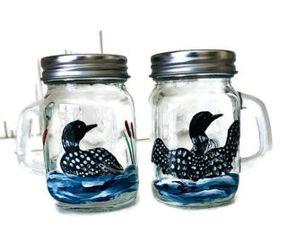 May include: Two glass mason jars with metal lids and handles. Each jar is painted with a loon, a type of bird, in black and white with a blue and white water scene.