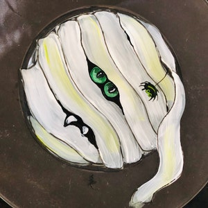 May include: A white and yellow painted mummy with green eyes and a black spider on a dark brown background.