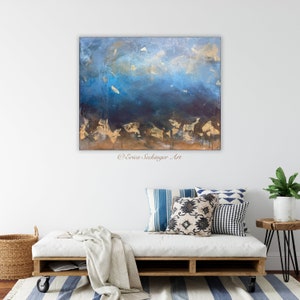 May include: Abstract painting with blue, gold, and brown hues. The painting features a textured, layered effect with gold accents. The painting is displayed on a white wall above a white couch with blue and white patterned pillows.