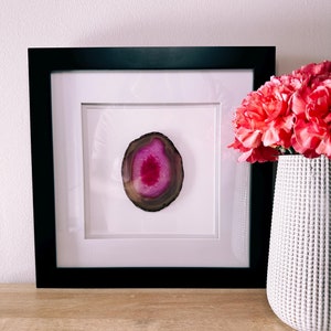 May include: A framed agate slice with a vibrant pink center and brown outer edges. The artwork is set against a white mat and framed in a black rectangular frame. A white textured vase with pink flowers is to the right.