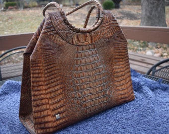 May include: A brown leather tote bag with a crocodile embossed pattern. The bag has a top handle and a long strap.