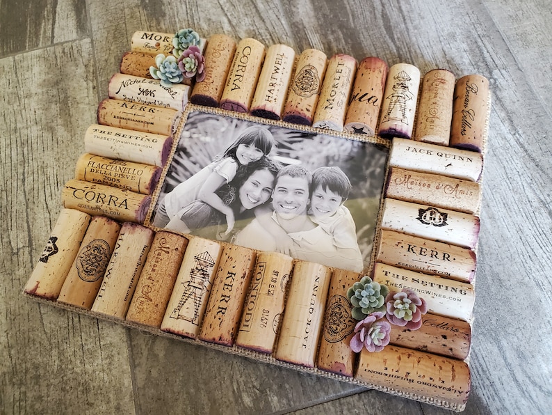 Puede incluir: Un marco de fotos hecho de corchos de vino, con una foto en blanco y negro de una familia de cuatro personas en su interior. El marco est&aacute; decorado con peque&ntilde;as suculentas verdes y rosas.