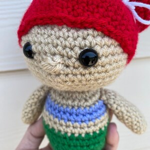 Op de afbeelding: Een gehaakte zeemeerminpop met een rode gehaakte hoed met een witte strik. De pop heeft bruin garenhaar, zwarte ogen en een groene staart met een blauwe en beige streep.