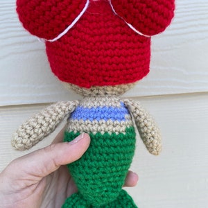 Op de afbeelding: Een gehaakte zeemeerminpop met rood haar in twee knotjes, een beige torso, een blauw en paars gestreept topje en een groene staart.