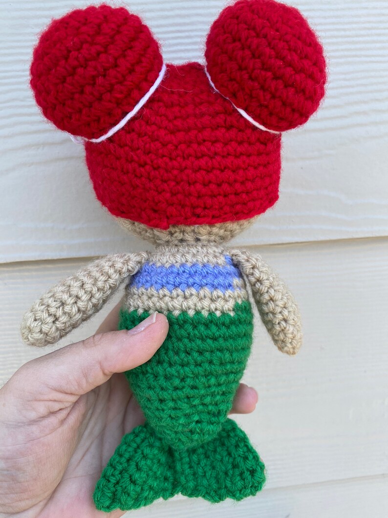 Op de afbeelding: Een gehaakte zeemeerminpop met rood haar in twee knotjes, een beige torso, een blauw en paars gestreept topje en een groene staart.