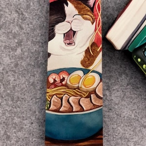 May include: A rectangular bookmark featuring a watercolor illustration of a calico cat with its mouth open, positioned above a blue bowl of ramen. The ramen includes noodles, two halved soft-boiled eggs, shrimp, and slices of pork belly.