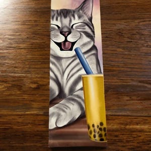 May include: A rectangular bookmark featuring a grayscale illustration of a smiling tabby cat with its eyes closed. The cat is positioned next to a yellow cup of bubble tea with dark brown tapioca pearls at the bottom and a blue straw sticking out.