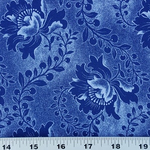 May include: Close-up of a blue fabric with a floral pattern. Large, stylized flowers and leafy vines are depicted in shades of blue and white. The background has a speckled texture. A ruler is at the bottom, with measurements in inches.