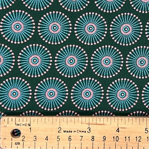 May include: A dark green fabric featuring a repeating pattern of circular motifs. Each circle consists of a central light blue ring surrounded by radiating light blue lines and a border of small pink dots. A wooden ruler shows a 5-inch scale.
