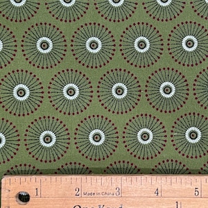 May include: Olive green fabric featuring a repeating pattern of circular sunburst motifs in black, white, and dark red. A wooden ruler is positioned at the bottom, showing measurements in inches, with the text "Made in China" and "One Yard".