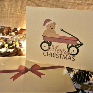May include: A Christmas card with a brown teddy bear wearing a Santa hat riding in a red wagon. The card has the text "Merry Christmas" on it.