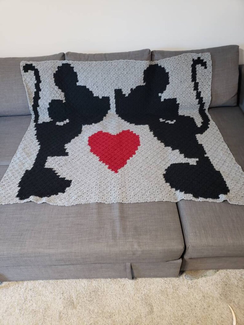 Puede incluir: Una manta de ganchillo con dos siluetas negras de gatos que se miran con un coraz&oacute;n rojo en el medio. La manta est&aacute; hecha con hilo gris y negro.