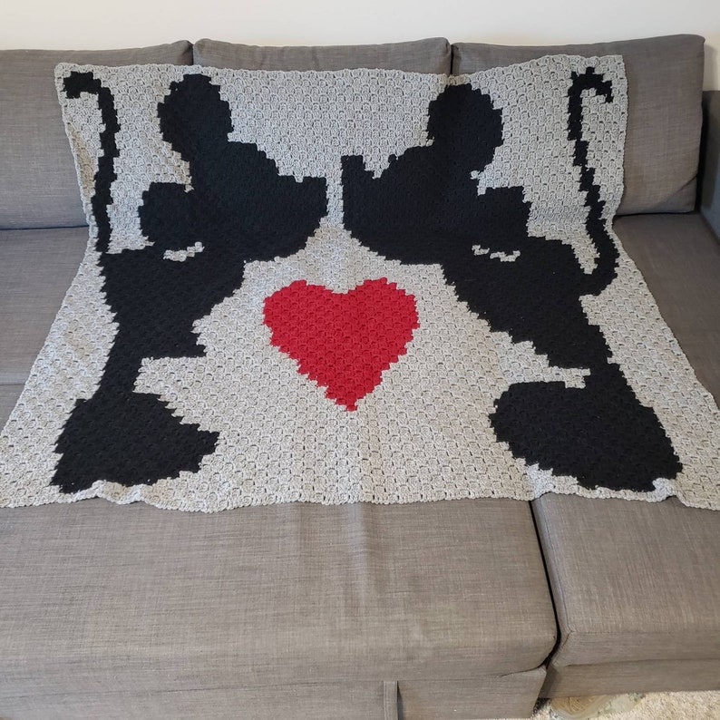 Puede incluir: Una manta de crochet gris con dos siluetas negras de personajes de dibujos animados que se toman de la mano y un coraz&oacute;n rojo en el centro.