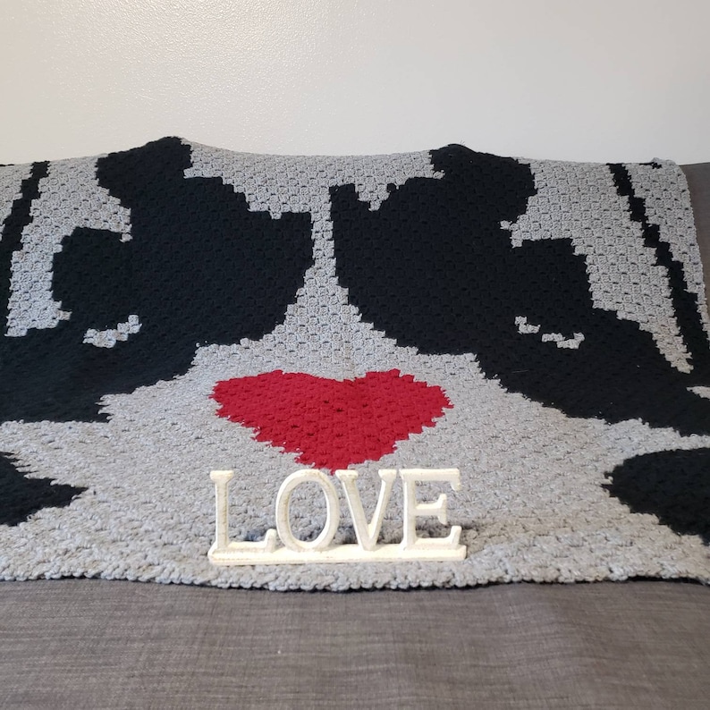 Puede incluir: Una manta de crochet gris y negro con un coraz&oacute;n rojo en el centro. La palabra "LOVE" est&aacute; escrita en letras blancas sobre un soporte delante de la manta.