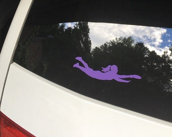 May include: A purple vinyl decal of a person diving, applied to a car window. The silhouette shows a person with outstretched arms and legs, in a diving pose. The background shows trees and a cloudy sky.