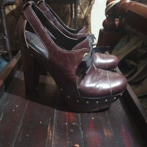 May include: A pair of brown leather platform heels with a black bow and gold studs. The heels are on a wooden surface.