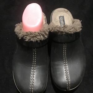 May include: A pair of black leather clogs with a brown faux fur lining. The clogs have a stitched seam down the front and the brand name "Crocs" is visible on the inside of one clog.