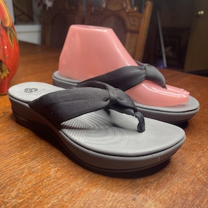 May include: A pair of black thong sandals featuring a grey footbed with a subtle circular pattern and a thick, soft fabric strap with a central knot detail. The sandals have a grey platform sole and a logo printed on the heel area of the footbed.