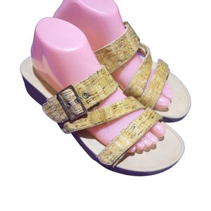 May include: A pair of beige sandals with cork-like straps and a buckle closure. The sandals have a light-colored sole and are displayed on pink foot forms. The straps are a light brown color.