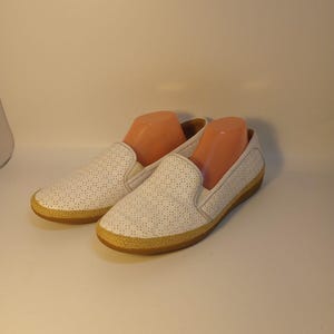 May include: White slip-on shoes with a perforated design. The shoes have a tan sole and a woven trim around the base. The shoes are displayed with shoe forms inside. These casual shoes are suitable for everyday wear.