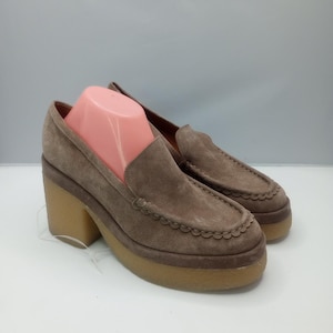 May include: A pair of taupe suede loafers featuring a thick platform sole and a chunky block heel. The shoes have a moccasin-style stitched toe detail with a scalloped edge trim along the vamp and a smooth, rounded silhouette throughout.