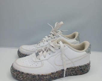 May include: White leather sneakers with a multi-colored speckled sole. The shoes have white laces, a Nike swoosh, and a padded collar. The sneakers are on a white surface.