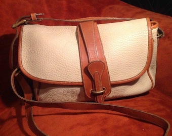 May include: Cream-colored leather shoulder bag with brown leather trim and a long adjustable strap. The bag features a flap closure with a brown leather strap and buckle. The bag is set against an orange background.