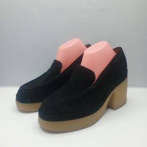 May include: A pair of black suede platform loafers with a rounded toe and a light brown sole. The shoes have a block heel and are displayed on pink shoe forms.