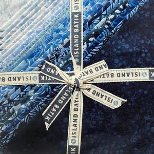 May include: A bundle of blue fabric squares, varying in shades from light to dark, tied with a ribbon that reads "ISLAND BATIK". The fabric has various patterns and textures, creating a visually appealing arrangement.