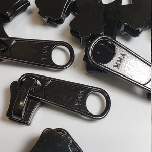 May include: A close-up of a pile of black metal zipper pulls. The zipper pulls are all the same size and shape, and they have a small loop at the top. The zipper pulls are all marked with the word "YKK".