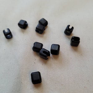 Puede incluir: Diez botones de pl&aacute;stico negro, cada uno con forma cuadrada y una peque&ntilde;a abertura en un lado. Los botones est&aacute;n esparcidos sobre una superficie de tela blanca.