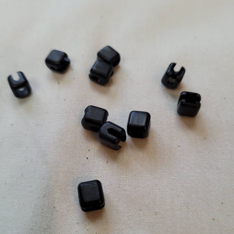 Puede incluir: Diez botones de pl&aacute;stico negro, cada uno con forma cuadrada y una peque&ntilde;a abertura en un lado. Los botones est&aacute;n esparcidos sobre una superficie de tela blanca.